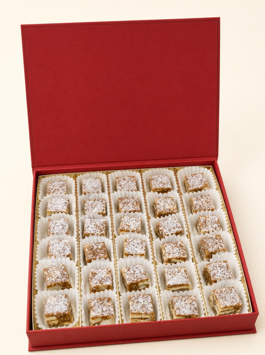 An open red Pure Red Gold gift box holding thirty coconut-dusted macadamia date squares in fluted white paper cups, on warm ivory