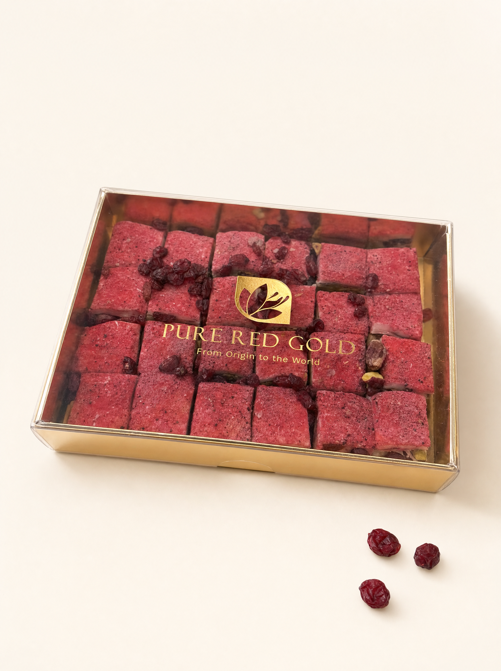 Gold gift box with a clear lid holding rose-red Signature Red Berry delights, the PURE RED GOLD lockup printed in gold on the lid, on warm ivory