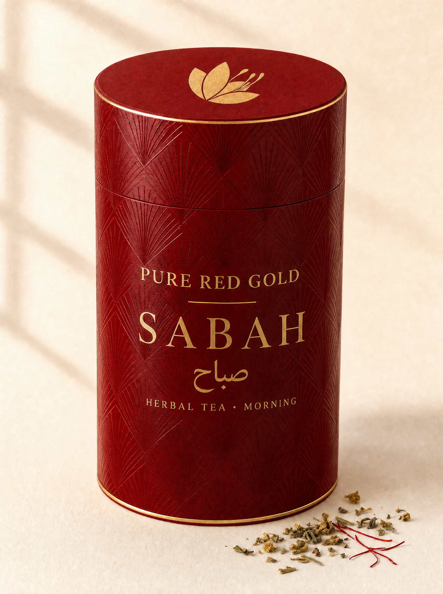 Sabah herbal tea — a crimson cylinder tube with a tone-on-tone diamond and fan pattern, gold foil rims, the saffron crocus on the lid and SABAH with its Arabic name in gold foil, on warm ivory